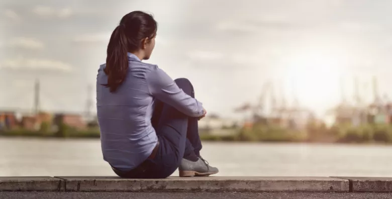 A woman sits facing away from us on the edge of a body of water and looks over.